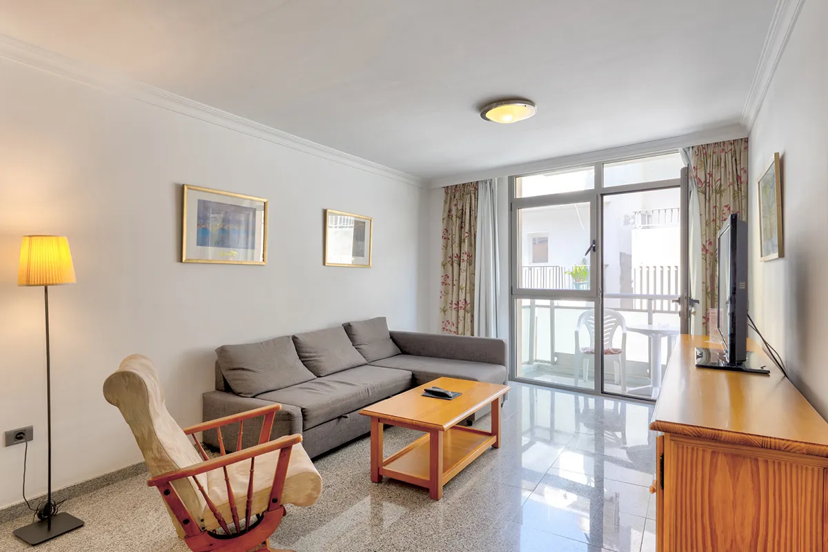 Sala de estar con sofá, silla, mesa de centro y TV junto a ventana amplia.