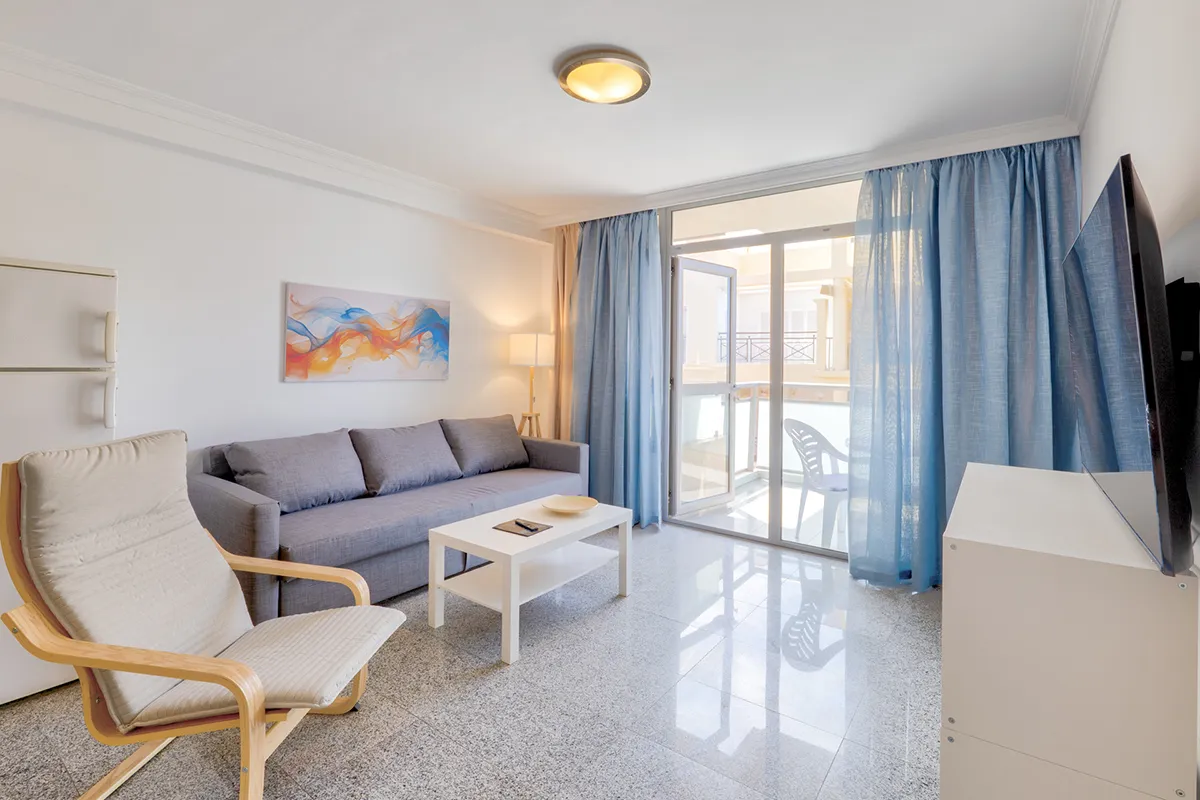 Sala luminosa con sofá gris, silla de madera y cortinas azules.