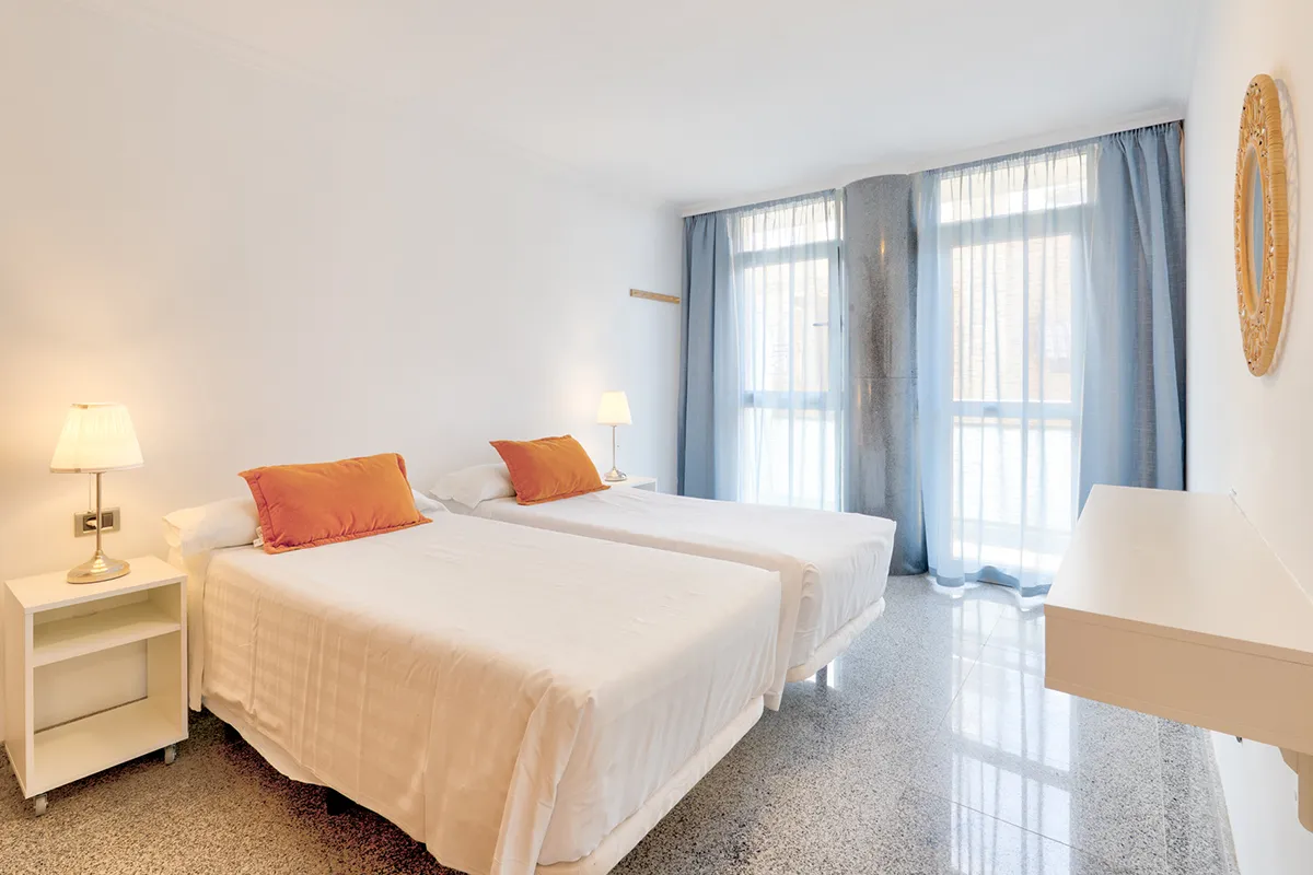 Habitación luminosa con dos camas, almohadas naranjas y luz de lámparas.