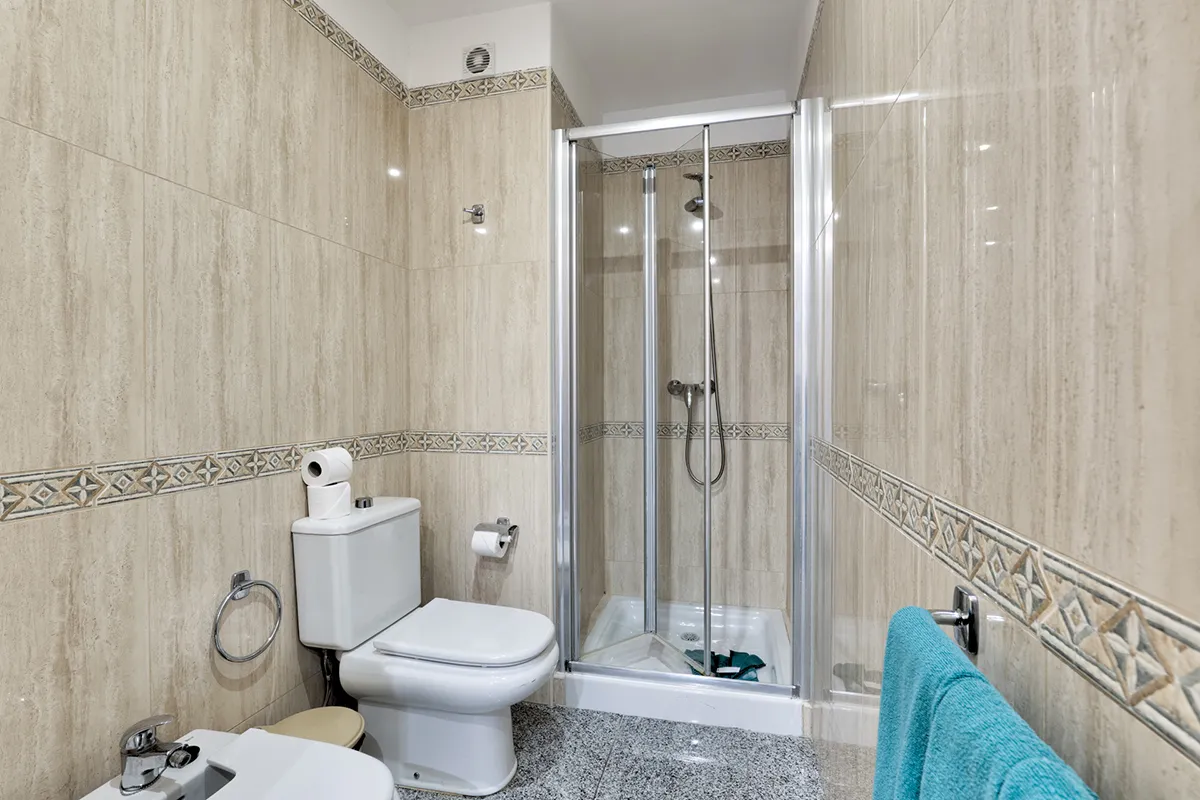 Baño con ducha, inodoro y bidé en azulejos beige y toalla azul.