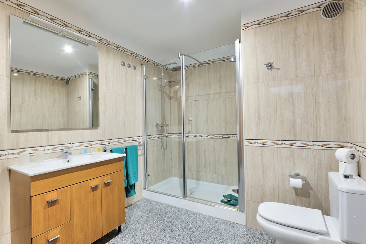 Baño moderno con ducha de vidrio, mueble de madera y azulejos beige.
