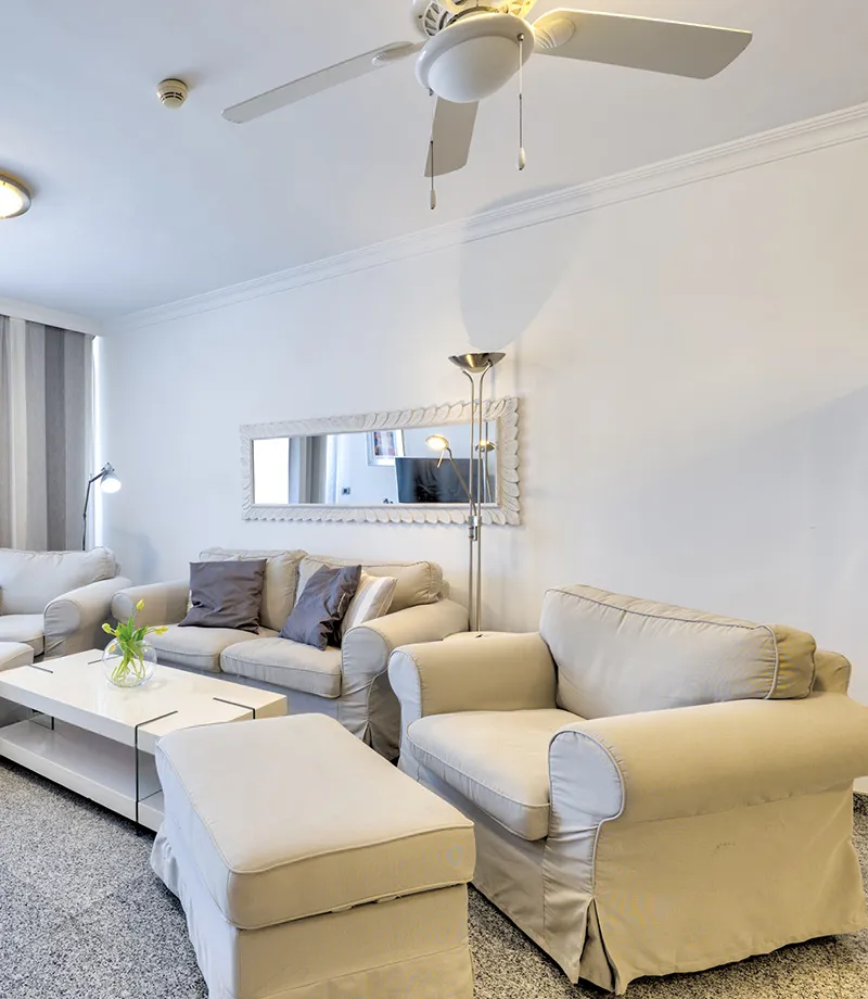 Sala acogedora con sof&aacute;s beige, espejo y ventilador de techo.