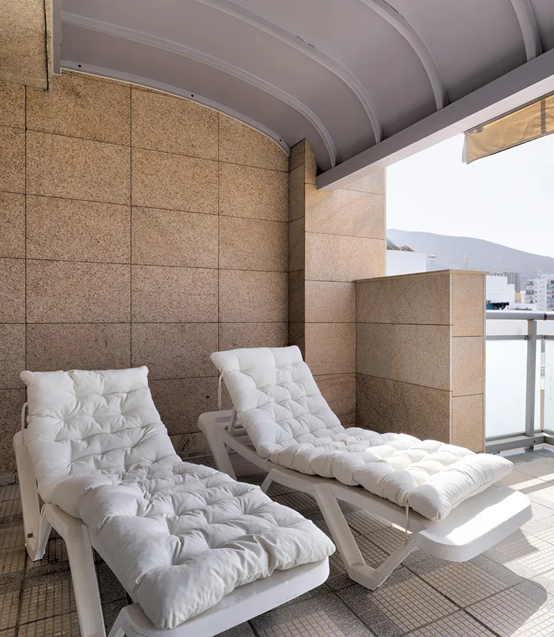 Camastros blancos bajo techo en un balc&oacute;n con paredes marrones.