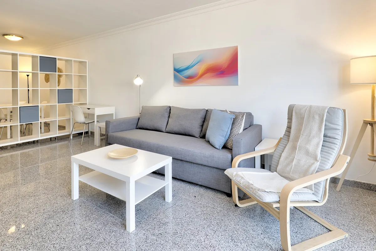 Sala moderna con sofá gris, mesa blanca y silla, iluminada con lámpara.