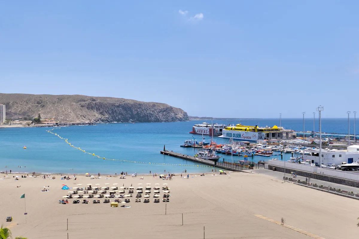 Playa con sombrillas y mar azul, fondo de montañas y puerto con ferris.