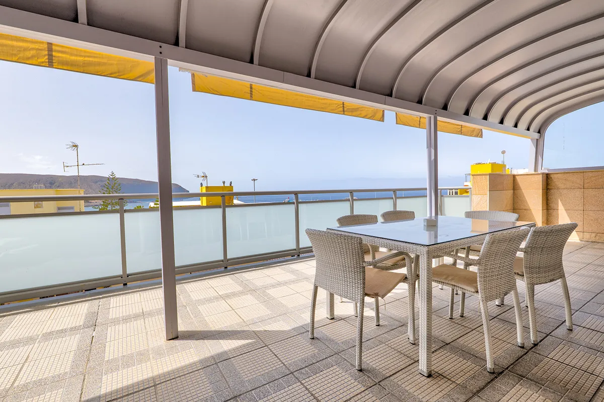 Terraza con mesa y sillas, vista al mar y montaña al fondo.