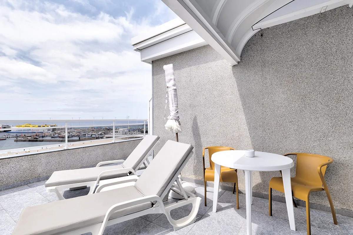 Terraza con vistas al mar, dos tumbonas, y mesa con sillas amarillas.