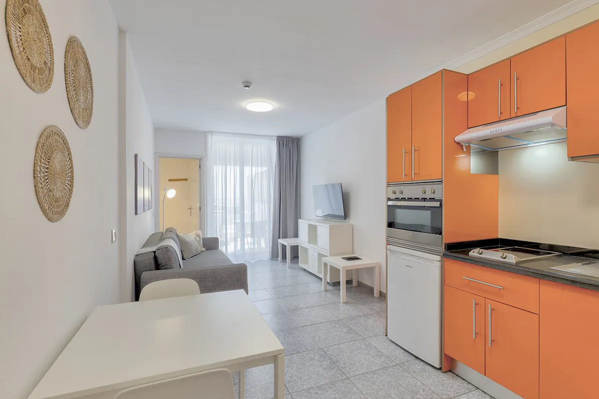 Sala moderna con cocina naranja a la derecha y sofá gris al fondo.