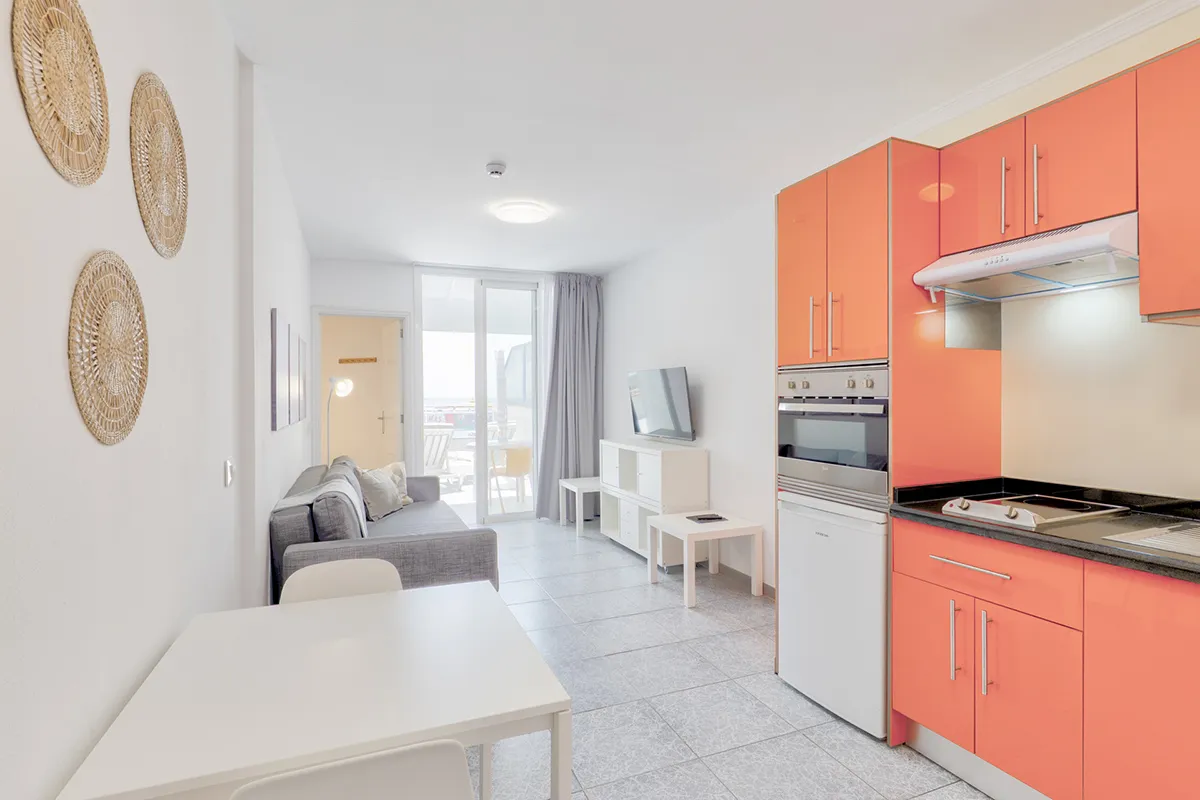 Sala moderna con cocina naranja, sofá, mesa y TV.