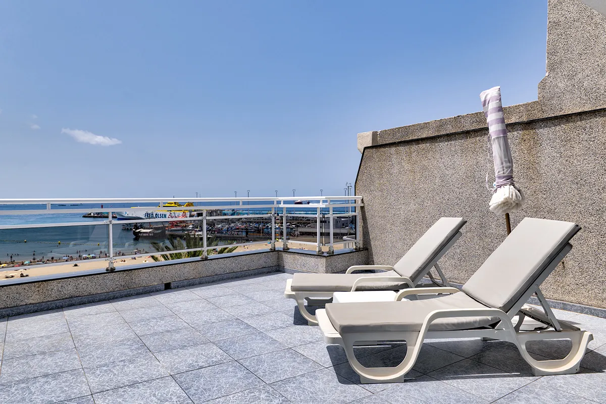 Terraza con tumbonas y vista al mar y muelle bajo un cielo azul despejado.