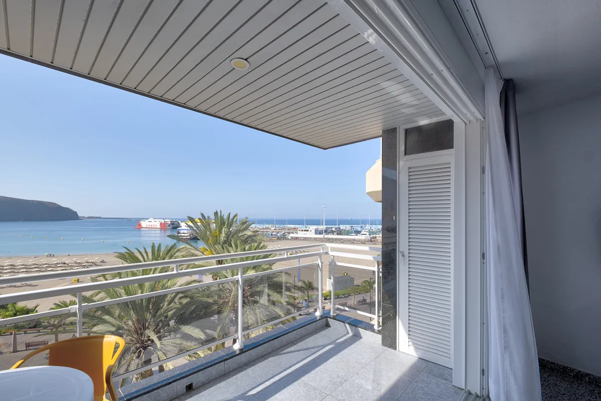 Vista desde un balcón hacia la playa, palmeras y un puerto con barcos.