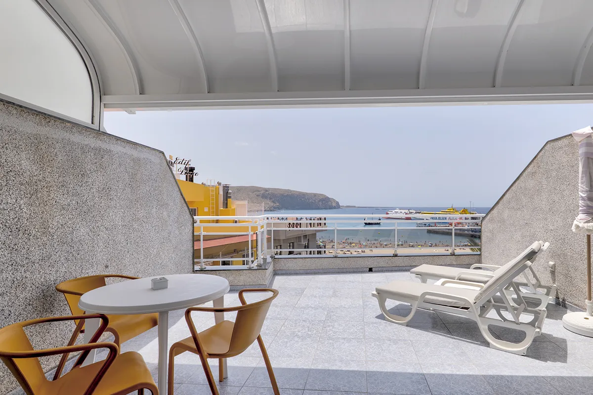 Balcón con mesa, sillas y vista al mar. Hay reposeras y barcos a la distancia.