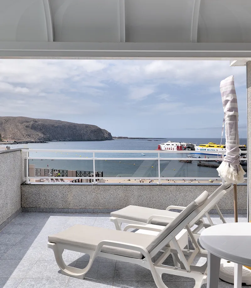 Terraza con tumbonas frente al mar y barcos en el puerto.