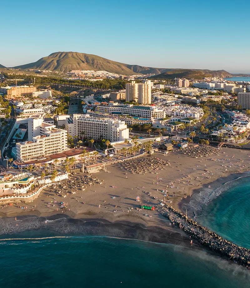 Vista a&eacute;rea de una playa con edificios y colinas al fondo.