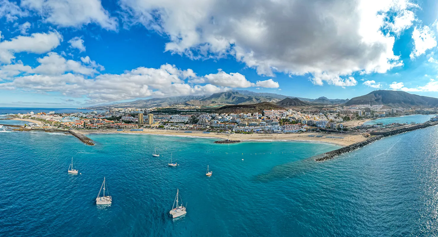 Vista a&eacute;rea de una ciudad costera con barcos y monta&ntilde;as al fondo.