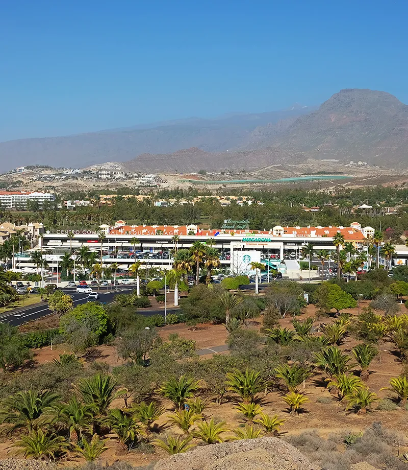 Centro comercial rodeado de palmeras, con monta&ntilde;as al fondo bajo un cielo azul.