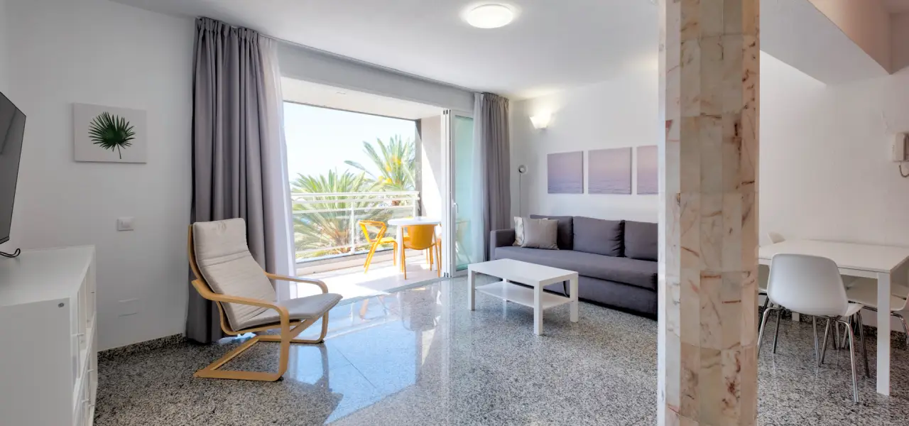 Sala moderna con sof&aacute;, mesa, silla y balc&oacute;n con vistas a palmeras.