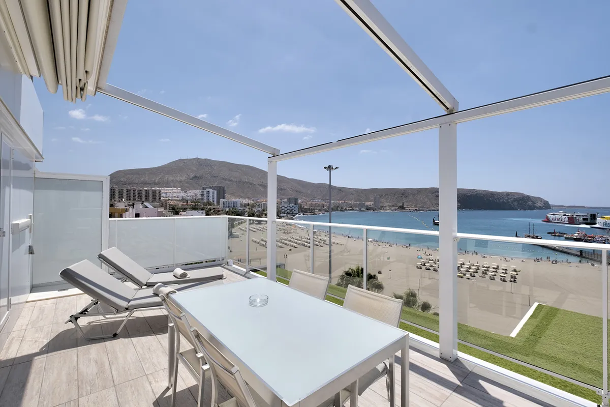 Terraza con vista al mar, tumbonas y mesa bajo pérgola en día soleado.