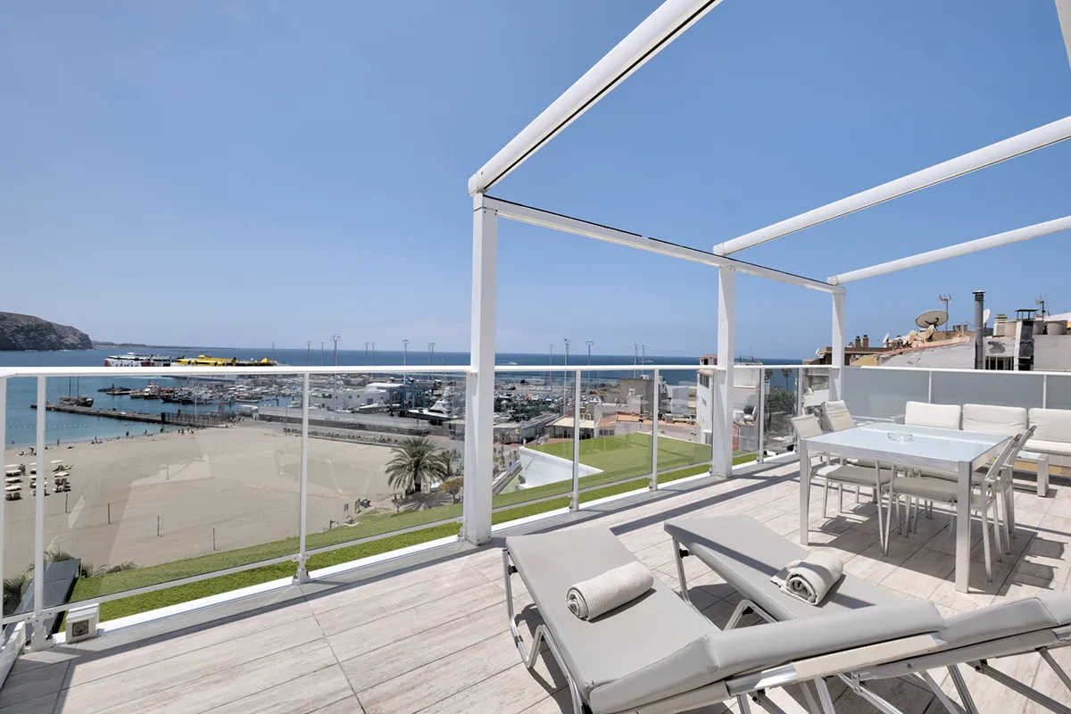Terraza con tumbonas y vista al mar con embarcaciones y playa.