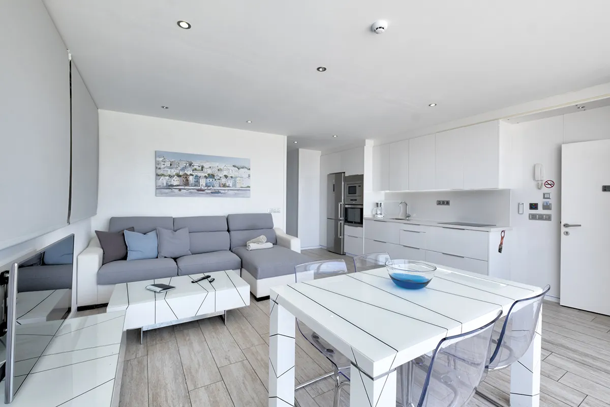 Sala y cocina modernas blancas con sofá gris y mesa con sillas transparentes.