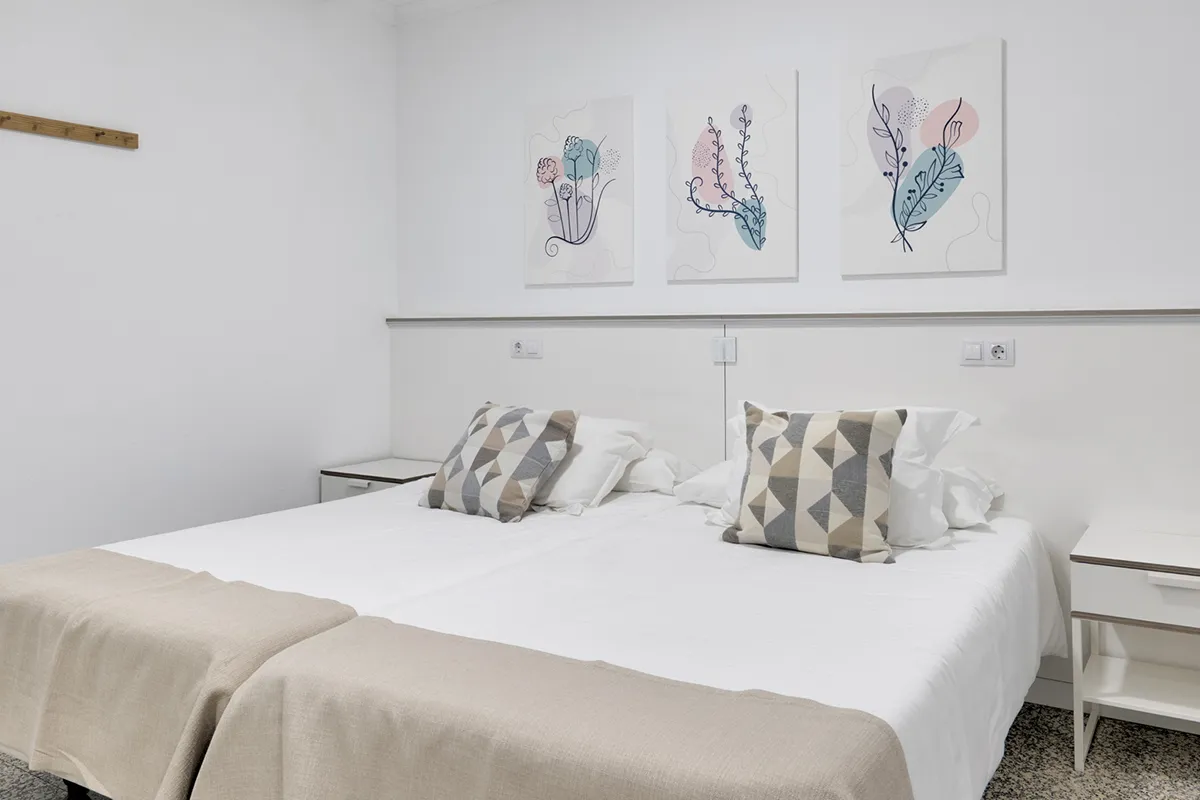 Habitación blanca con dos camas, cojines geométricos y cuadros de plantas.
