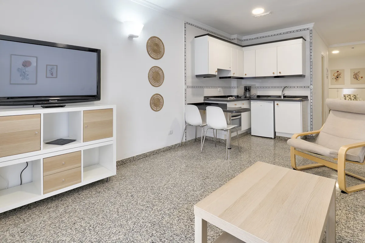 Sala y cocina integradas con muebles blancos, televisor y sillón beige.