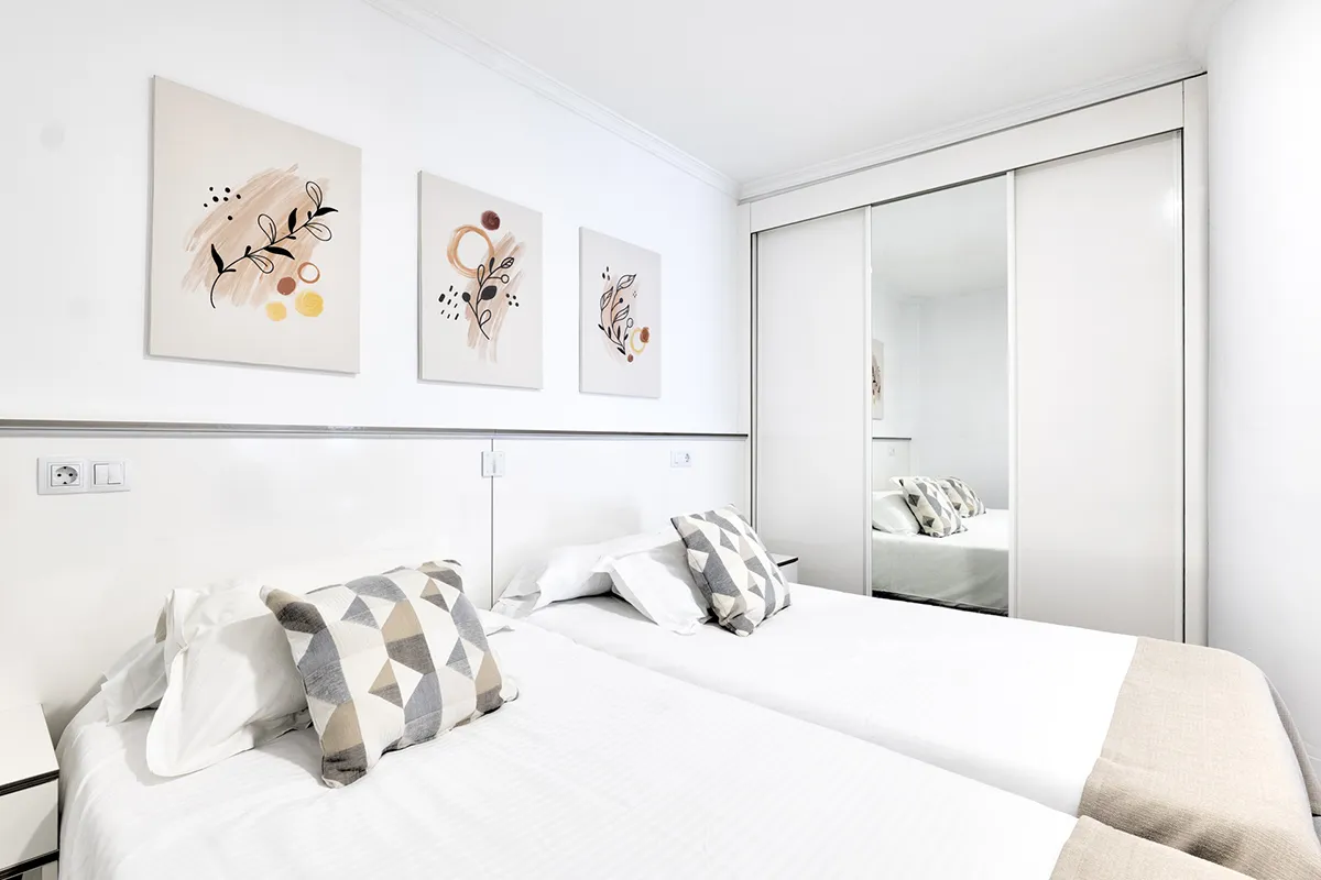 Habitación blanca con dos camas, cojines geométricos y cuadros abstractos.