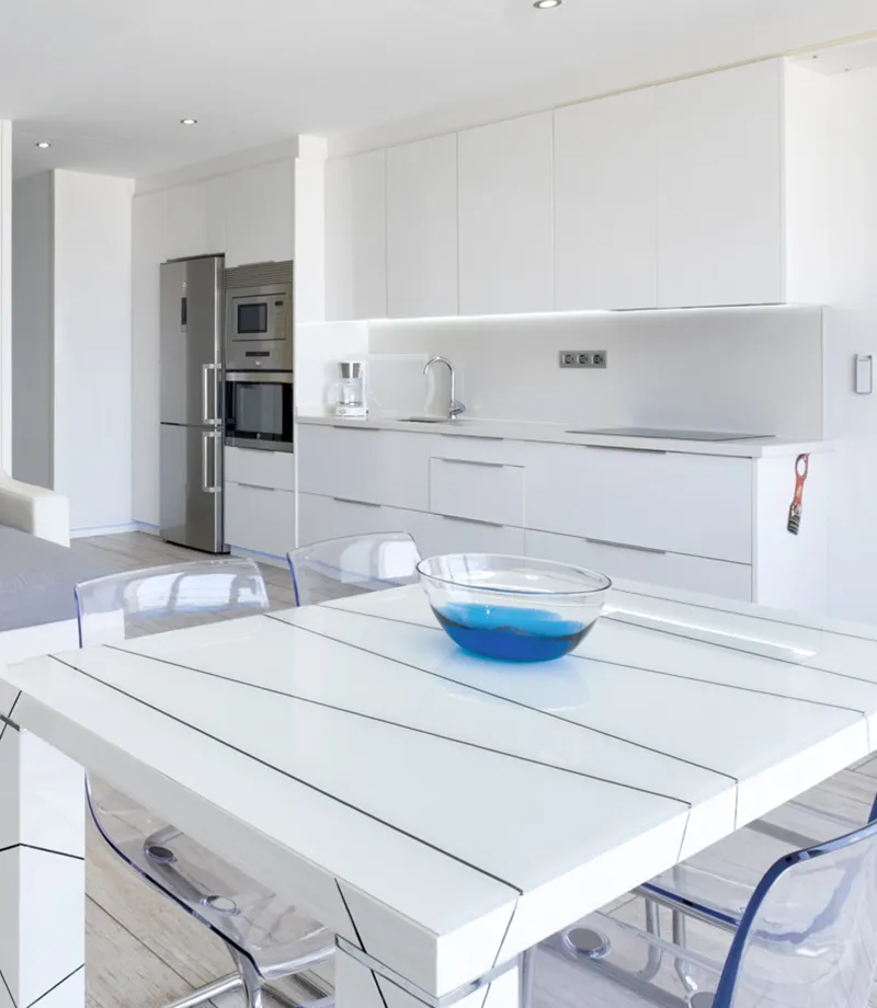 Cocina moderna blanca con mesa y sillas transparentes; taz&oacute;n azul en la mesa.