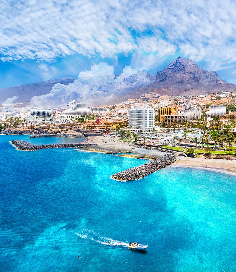 Costa con aguas turquesa, monta&ntilde;as al fondo y edificios modernos.