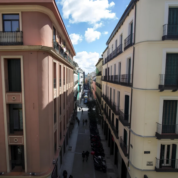 Calle estrecha entre edificios altos con peatones y motos estacionadas. Cielo despejado.