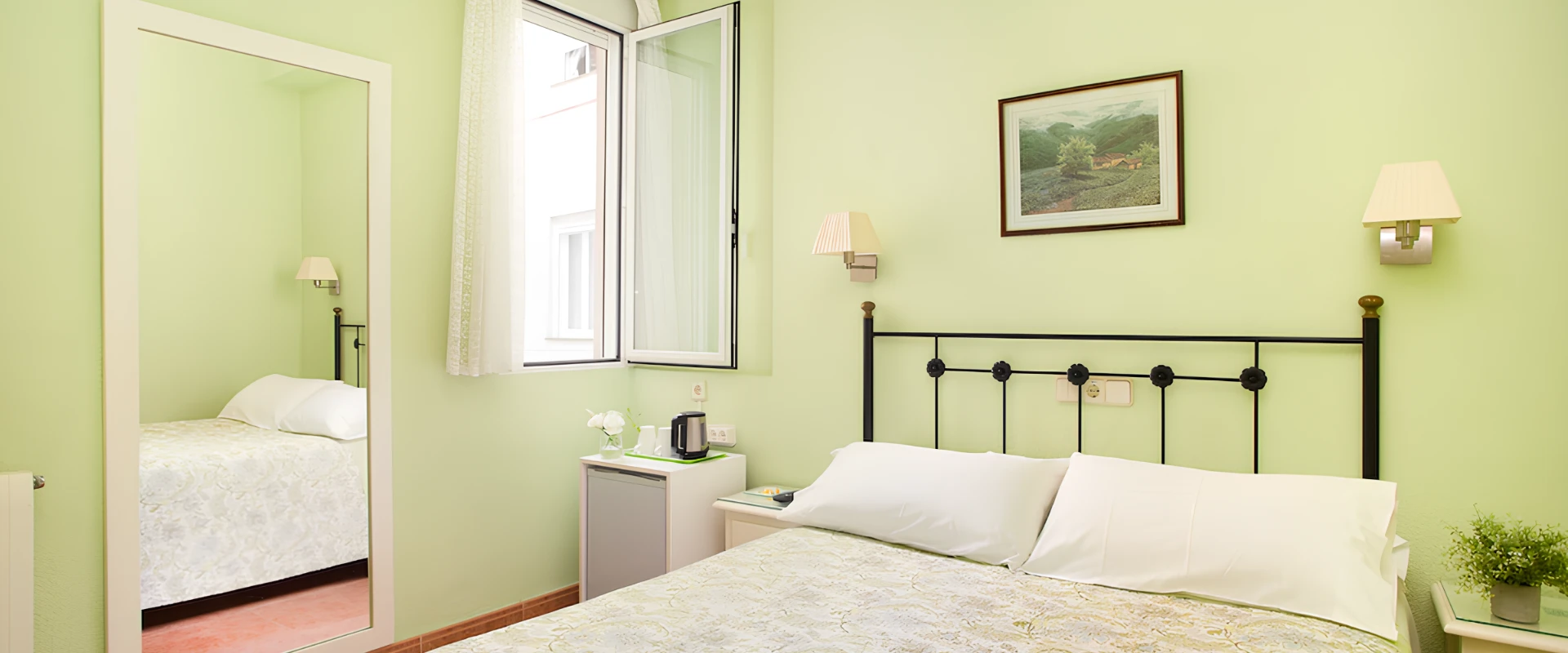 Dormitorio con paredes verdes, cama, l&aacute;mpara de pared, ventana abierta y espejo grande.