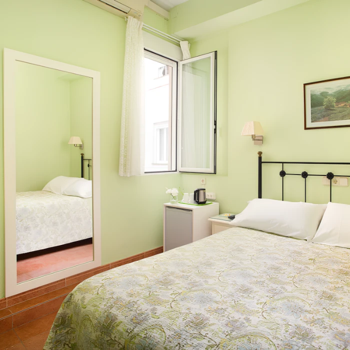 Habitaci&oacute;n luminosa con cama, espejo, ventana abierta, y mesita con tetera sobre mini nevera.