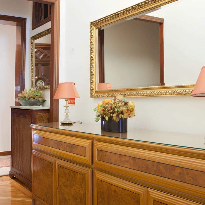Mueble de madera barnizada con espejo dorado, l&aacute;mpara y flores decorativas en una entrada elegante.