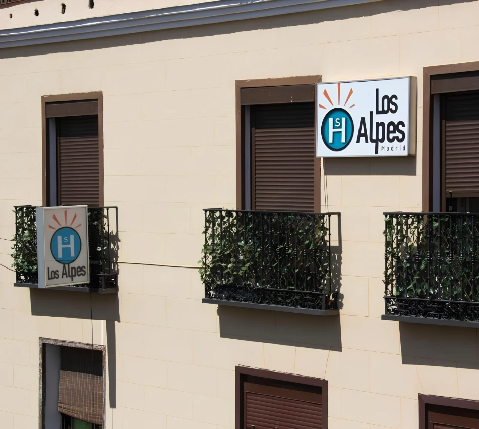 Edificio con dos carteles de "Los Alpes Madrid", ventanas y balcones con rejas y plantas.