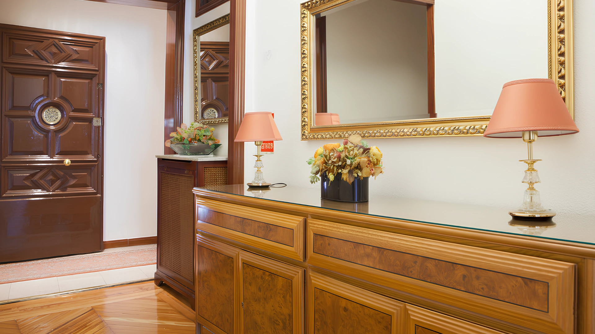 Entrada elegante con puerta de madera, espejo dorado, l&aacute;mparas y florero sobre una c&oacute;moda.