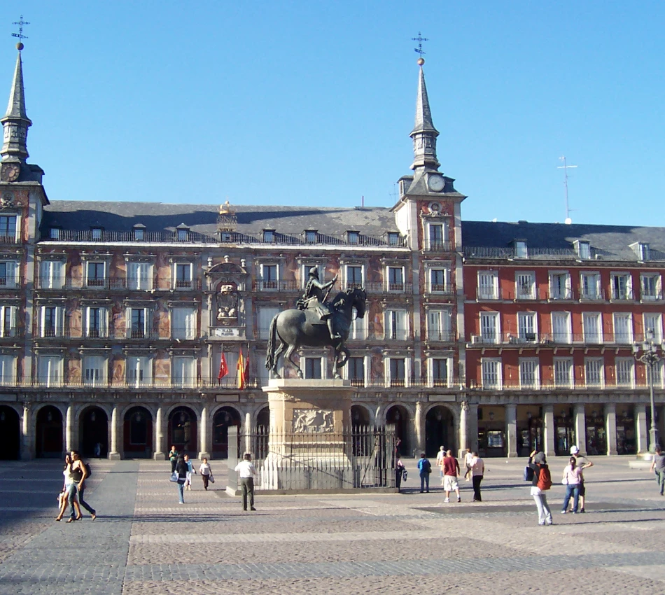 Plaza con edificio hist&oacute;rico, estatua ecuestre central y gente paseando; cielo despejado.