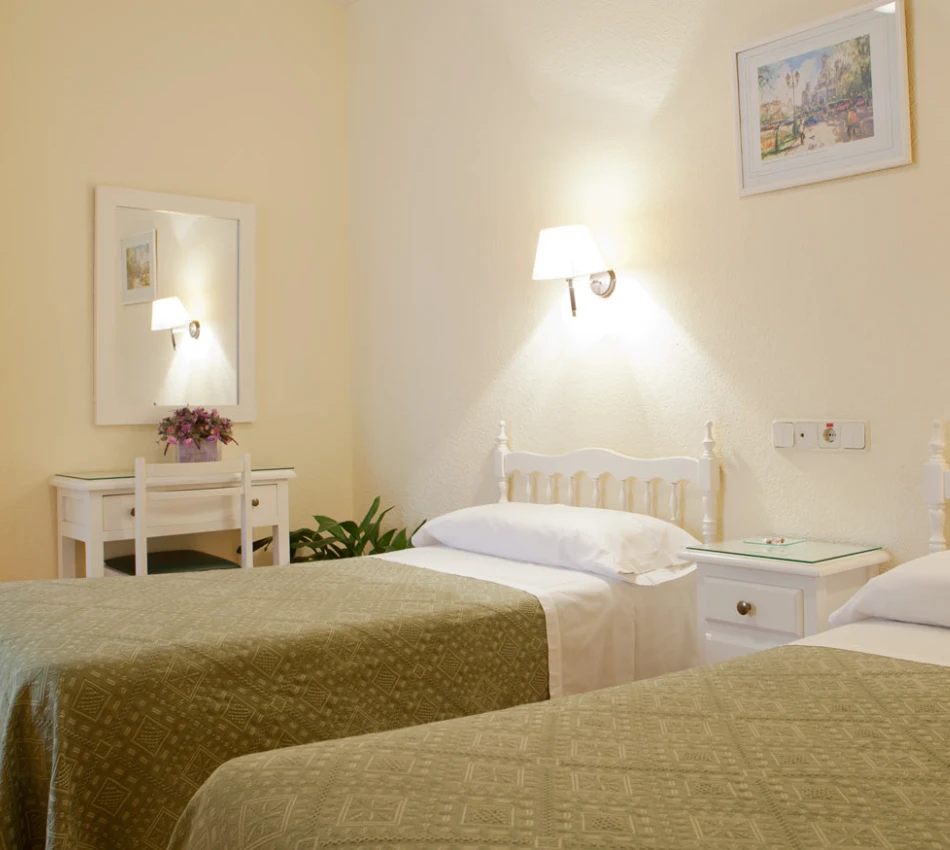 Habitaci&oacute;n con dos camas, mesa con espejo, l&aacute;mpara de pared y cuadro decorativo encima.