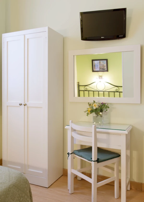 Habitaci&oacute;n con armario, espejo, televisor, escritorio, silla blanca y flores en un jarr&oacute;n.