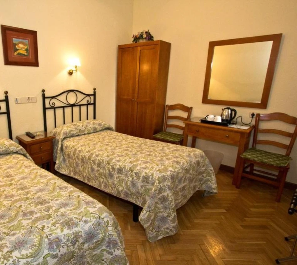 Habitaci&oacute;n sencilla con dos camas, armario, mesa con tetera, espejo y dos sillas de madera.