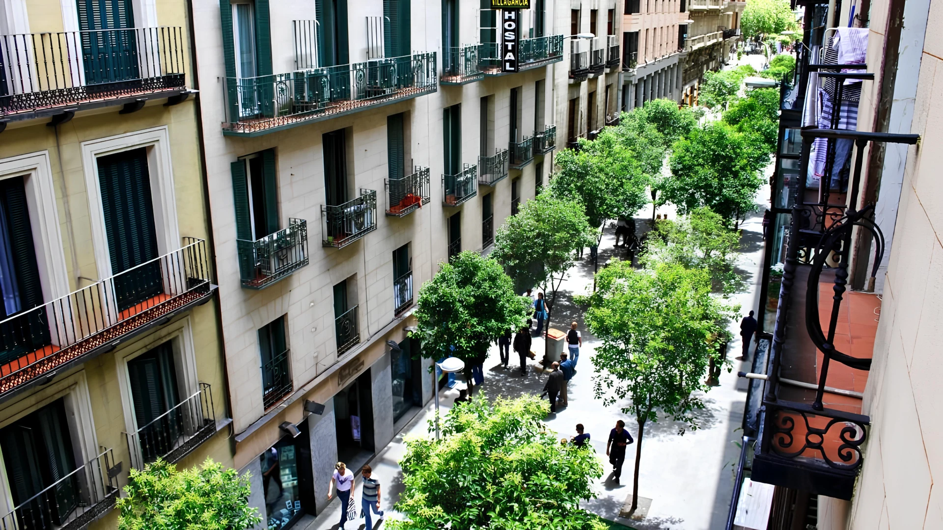 Calle peatonal arbolada con edificios altos y balcones. Personas caminando.