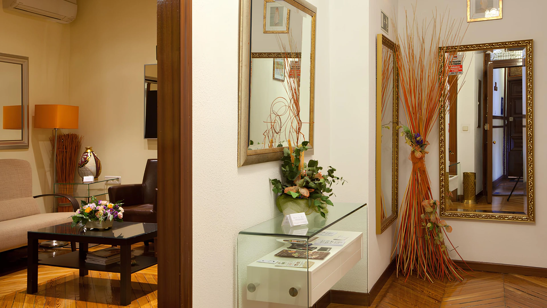Sala de espera elegante con espejos, mesa de vidrio y arreglos florales en la entrada.