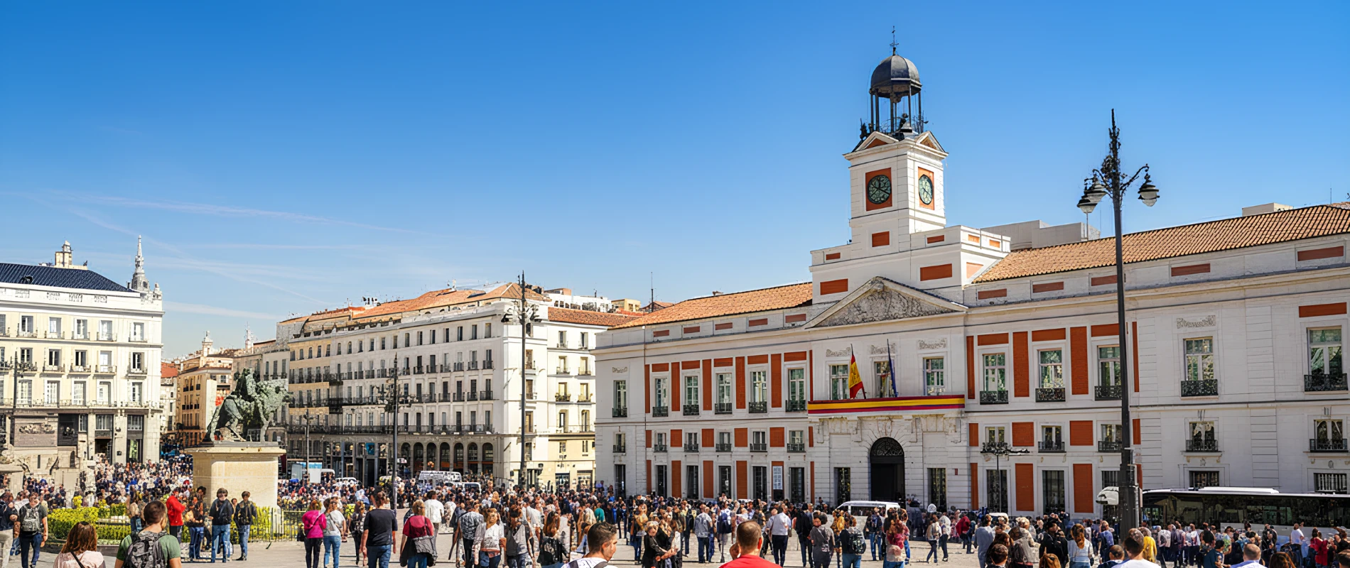 Multitud en una plaza con edificio hist&oacute;rico y reloj bajo un cielo azul.