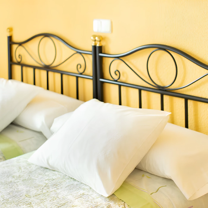 Cama con cabecero de metal negro y almohadas blancas sobre un fondo de pared amarilla.