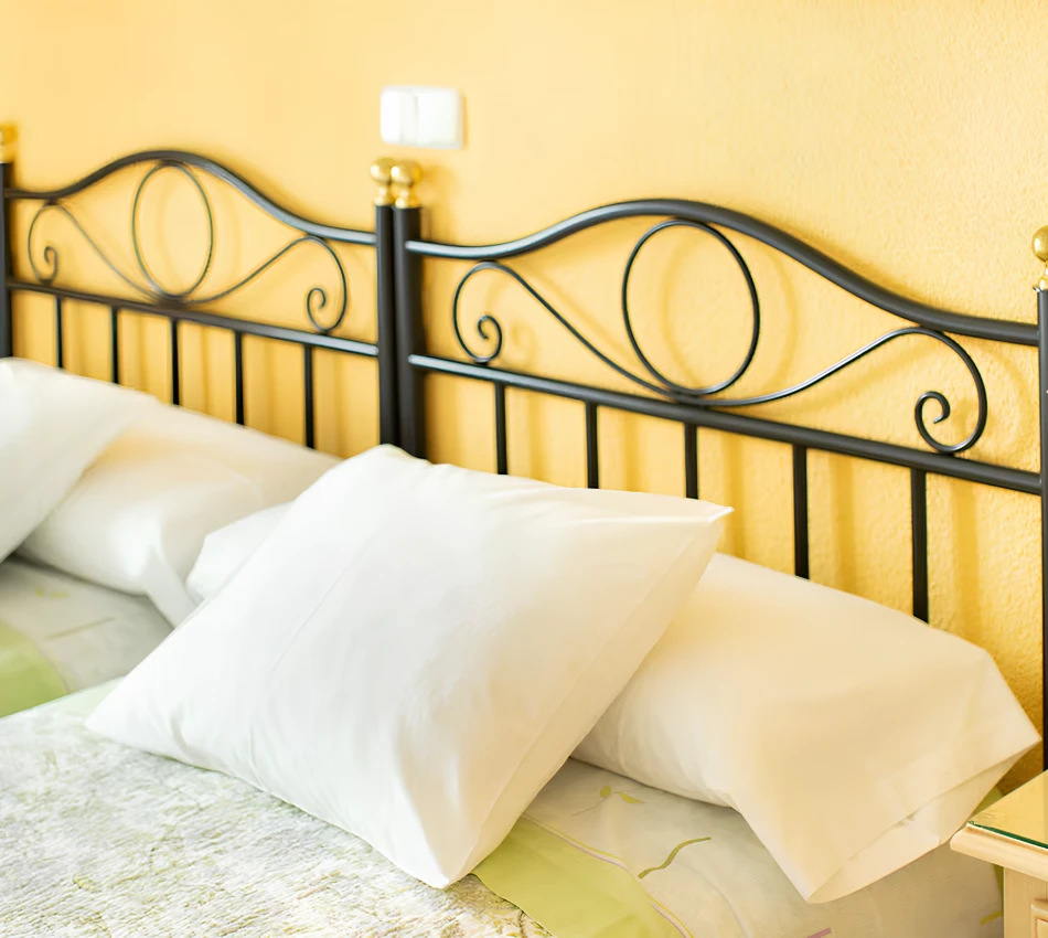 Cama con cabecero de metal negro y cojines blancos sobre colch&oacute;n, pared amarilla de fondo.