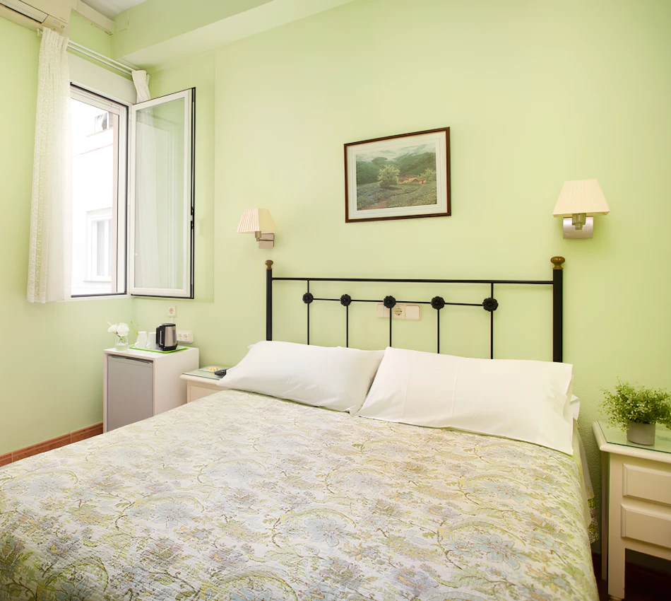 Habitaci&oacute;n luminosa con pared verde, cama doble, ventana abierta y decoraci&oacute;n sencilla.