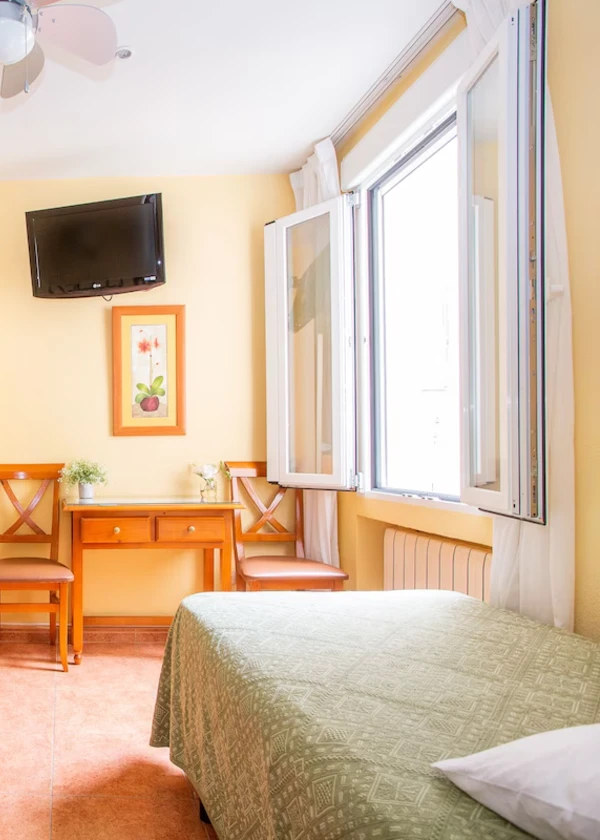 Habitaci&oacute;n luminosa con cama, dos sillas, escritorio, ventana abierta y televisi&oacute;n en la pared.