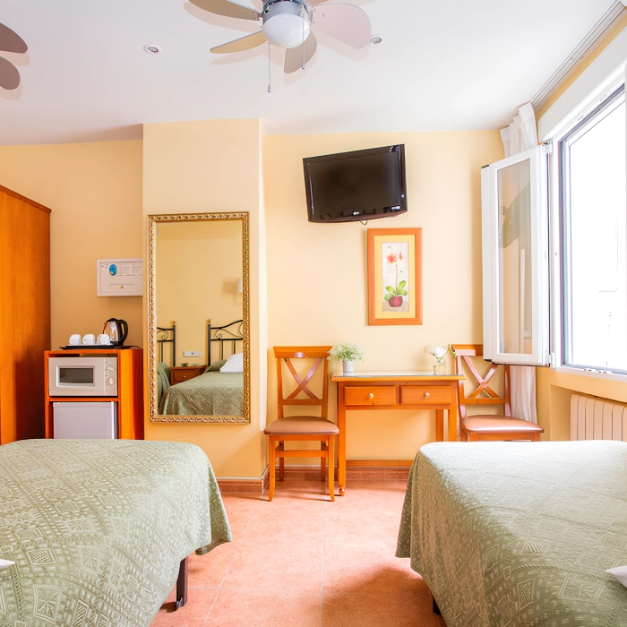 Habitaci&oacute;n con dos camas, TV en la pared, escritorio con sillas, ventana abierta y ventilador.