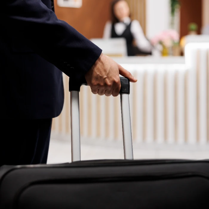 Persona con equipaje en recepci&oacute;n de hotel, fondo con recepcionista desenfocada.