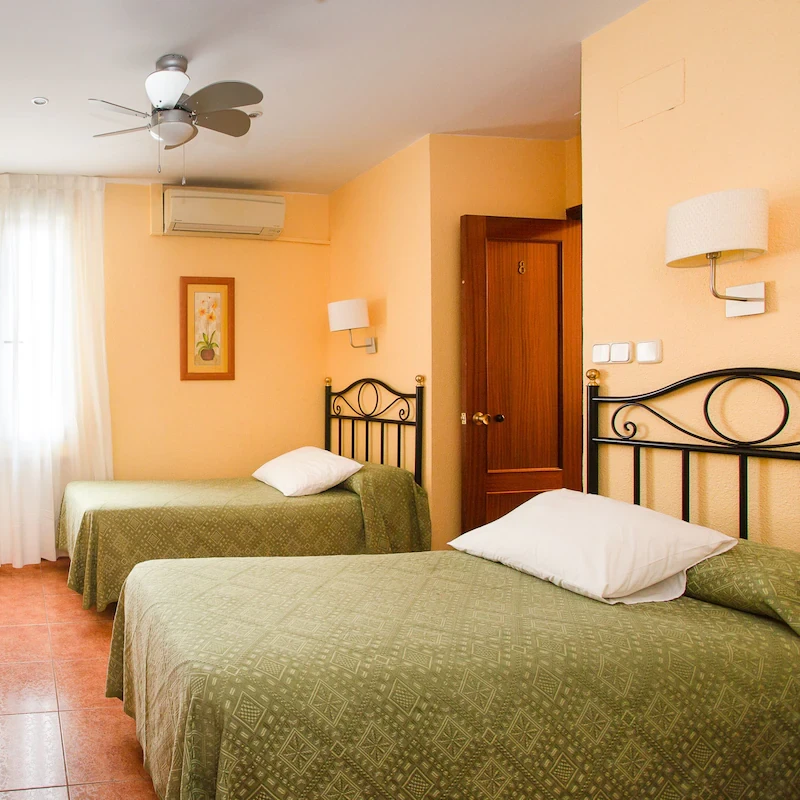 Habitaci&oacute;n con dos camas individuales, paredes beige, colchas verdes y ventilador de techo.