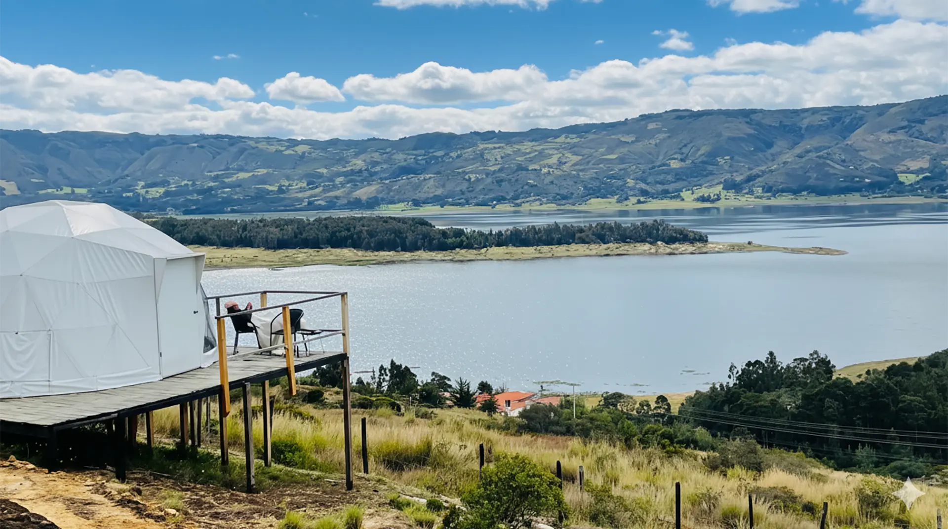 Persona descansando junto a tienda domo cerca de un lago en un paisaje monta&ntilde;oso.